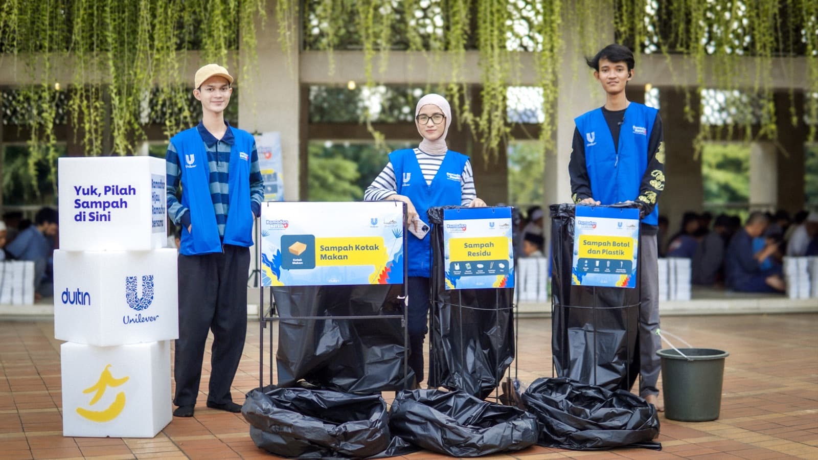 Three volunteers in blue Unilever vests standing behind Duitin waste-sorting bins labelled for food boxes, residue, and bottles and plastic
