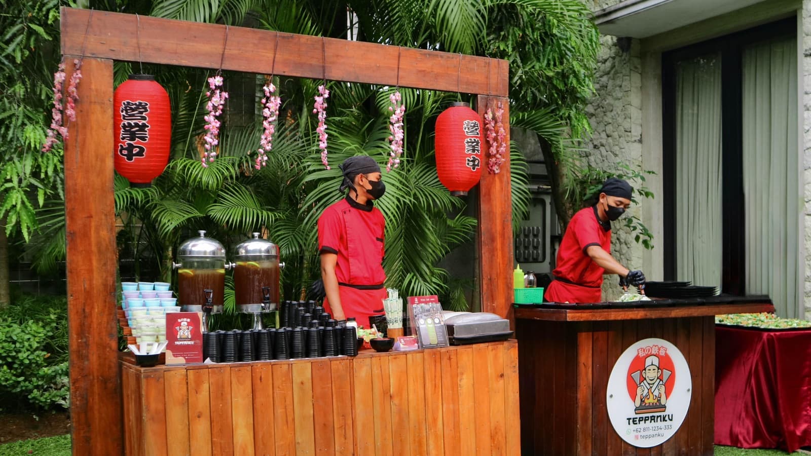 Two chefs in red uniforms at a wooden Teppanku catering stall under red paper lanterns, in a garden